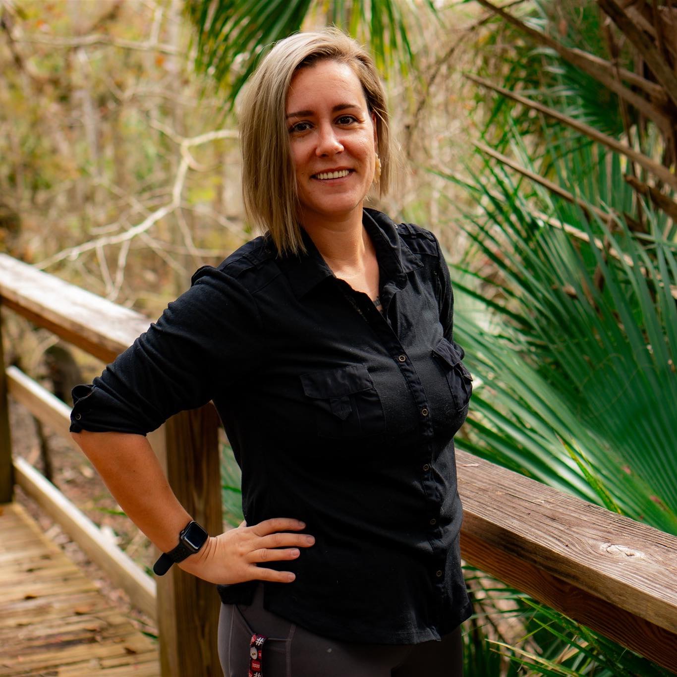 A woman with short blonde hair, wearing a black button-up shirt and a smartwatch, stands smiling on a wooden bridge surrounded by lush green foliage.