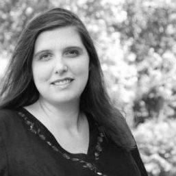 Black and white photo of a woman with long hair, wearing a dark top, standing outdoors with trees and foliage blurred in the background. She is looking at the camera and smiling slightly.