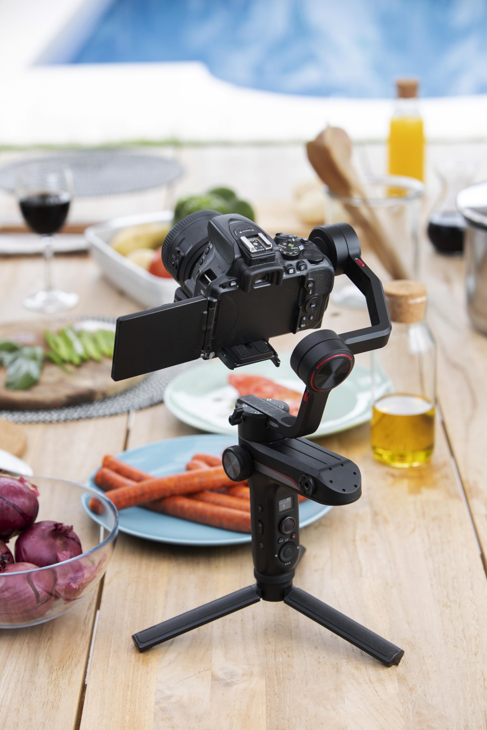 A digital camera mounted on a gimbal tripod sits on a wooden table with plates of vegetables, bottles, and glassware in the background, near a swimming pool.